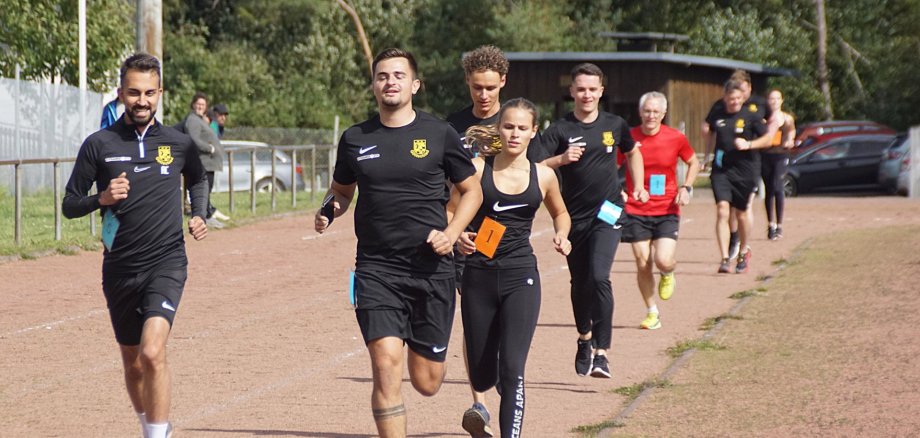 Das Foto zeigt neun Sportlerinnen und Sportler, die hintereinander auf einer Laufbahn laufen.