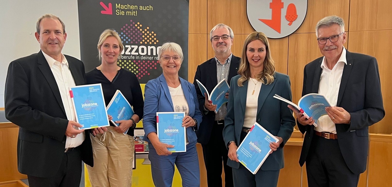 Das Foto zeigt die genannten Personen, die mit jeweils einer Broschüre in der Hand im Sitzungssaal stehen.