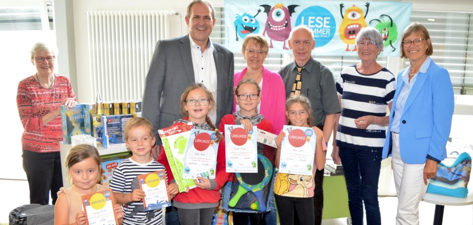 Das Foto zeigt die genannten Personen. Sie stehen in der Stadtbibliothek zusammen mit einigen Kinder, die ihre Urkunden in der Hand halten, vor einem Tisch mit Tombola-Gewinnen.