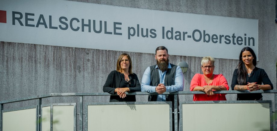 Das Foto zeigt die genannten Personen, die in einer Reihe vor einem Schriftzug mit der Aufschrift "Realschule plus Idar-Oberstein"  auf einem Geländer lehnen.