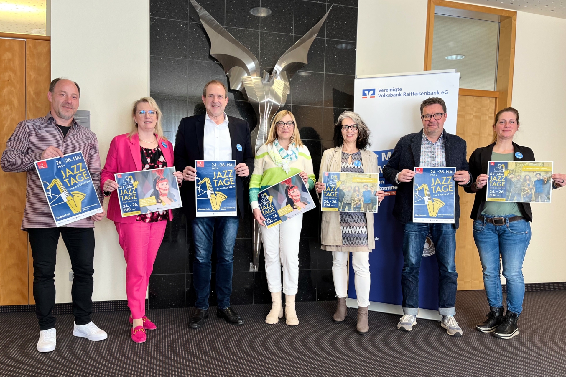Das Foto zeigt sieben Personen. Sie halten Plakate der Jazztage in der Hand, stehen nebeneinander vor einer Skulptur und schauen in die Kamera.