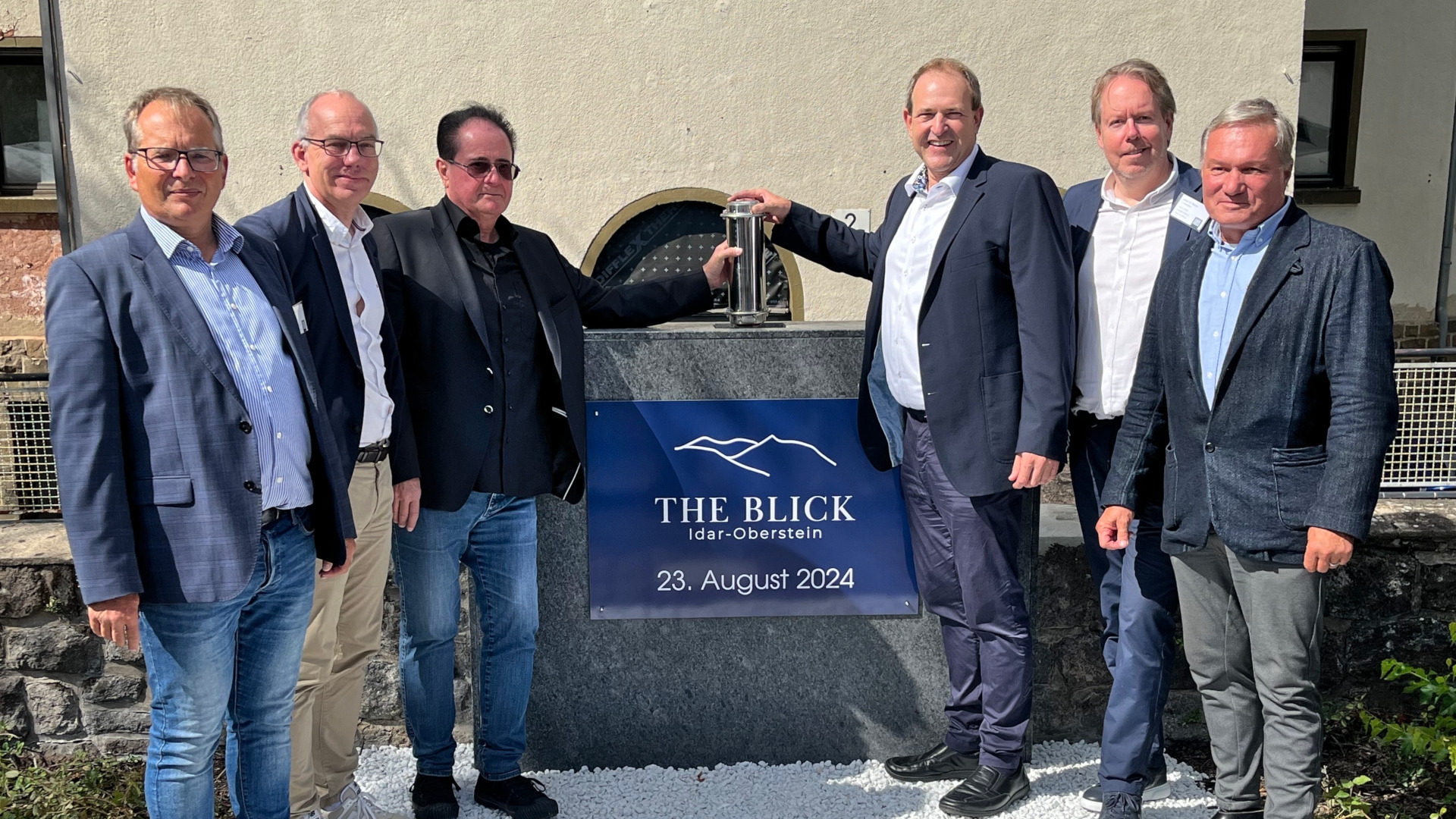 Das Foto zeigt die genannten Personen, die nebeneinander vor einem Granitblokc mit der Aufschrift "The Blick Idar-Oberstein - 23. August 2024" stehen. Herr Haferkorn und OB Frühauf halten die Zeitkapsel über das Loch, in das sie versenkt wird.