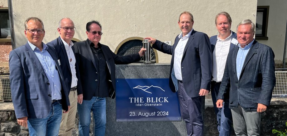 Das Foto zeigt die genannten Personen, die nebeneinander vor einem Granitblokc mit der Aufschrift "The Blick Idar-Oberstein - 23. August 2024" stehen. Herr Haferkorn und OB Frühauf halten die Zeitkapsel über das Loch, in das sie versenkt wird.