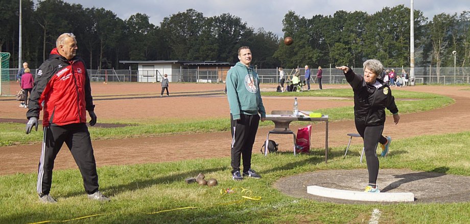Das Foto zeigt eine Frau im Trainingsanzug beim Kugelstoßen. Zwei Kamprichter sehen ihr zu. 