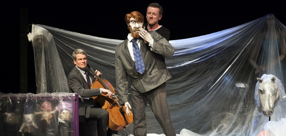 Das Foto zeigt einen Schauspieler mit einer lebensgroßen Figur auf einer Bühne. Hinter ihm sitzt ein Cellist auf einem Hocker.