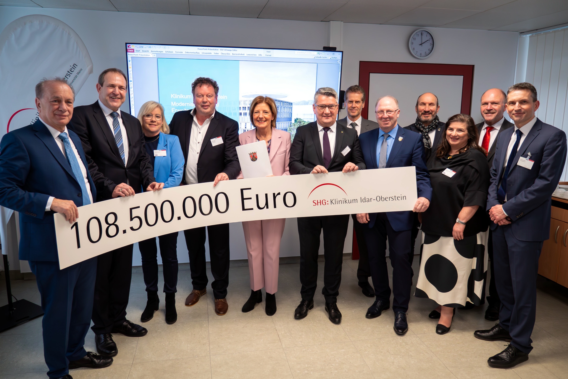 Das Foto zeigt die genannten Personen. Sie stehen nebeneinander in einem Besprechungsraum des Klinikums. Sie halten eine langes Transparent, auf dem der Betrag 108.500.000 Euro und das Logo des Klinikums Idar-Oberstein aufgedruckt sind, in den Händen.