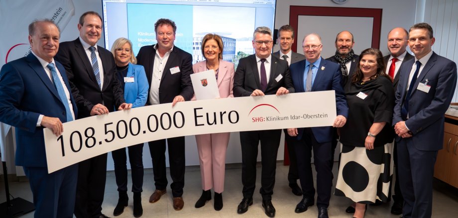 Das Foto zeigt die genannten Personen. Sie stehen nebeneinander in einem Besprechungsraum des Klinikums. Sie halten eine langes Transparent, auf dem der Betrag 108.500.000 Euro und das Logo des Klinikums Idar-Oberstein aufgedruckt sind, in den Händen.