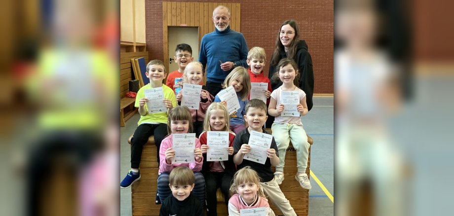 Das Foto zeigt elf Kinder, Klaus Juchem und eine Erzieherin. Die Kinder sitzen in der Sporthalle vor und auf einem Kasten (das Sportgerät) und halten Urkunden in den Händen. Die Erwachsenen stehen dahinter.