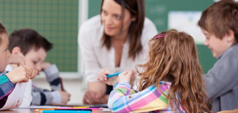 Das Foto zeigt eine Lehrerin und mehrere Schüler in einem Klassenraum. Die Schüler sitzen an einem Tisch, die Lehrerin beugt sich zu ihnen.