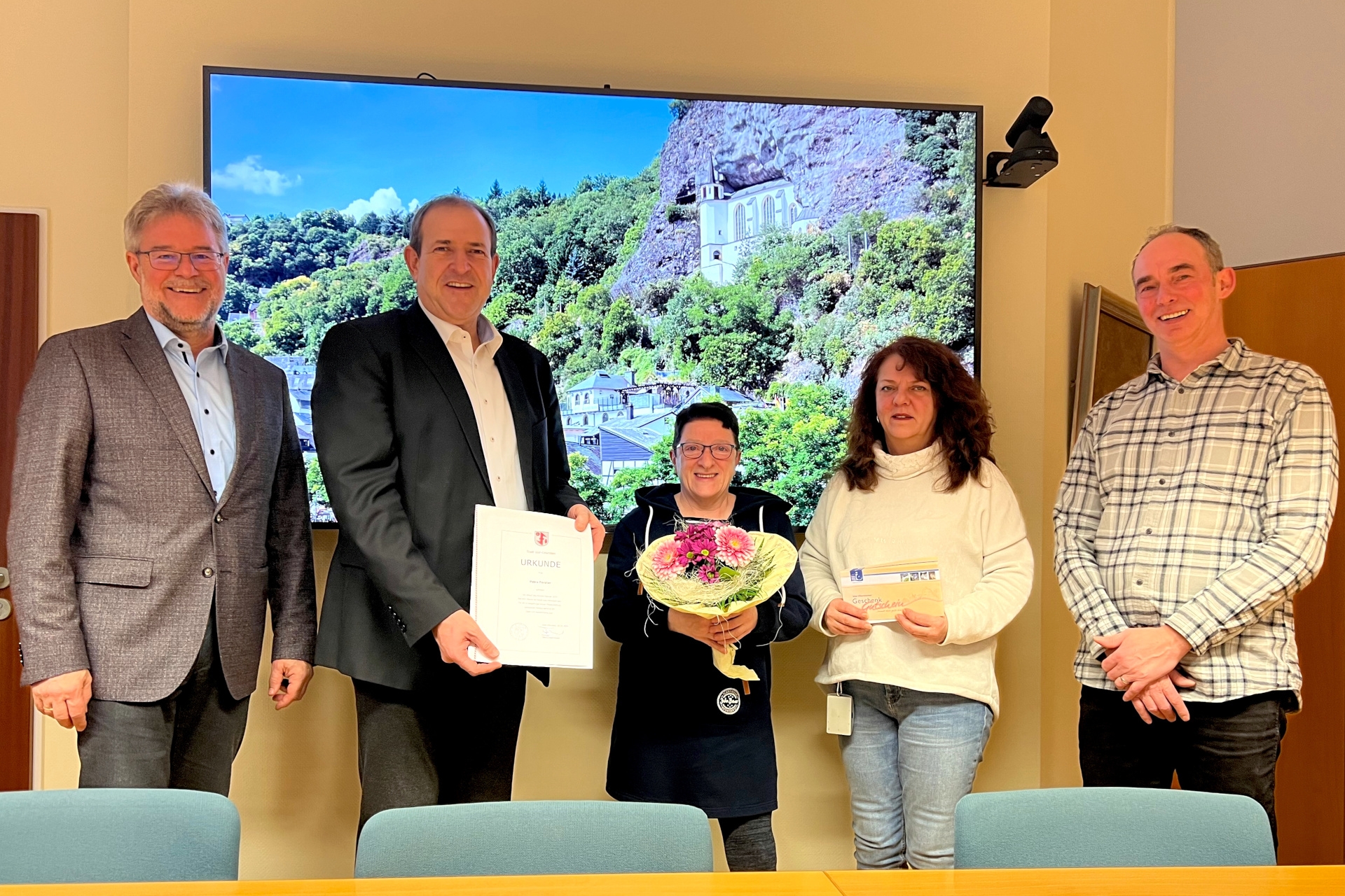 Auf dem Bild stehen der Leitende Bürobeamte Wolfgang Petry, OB Frank Frühauf, Petra Forster, Personalratsvorsitzende Susanne Becker und Jugendamtsleiter Michael Schweizer (v. l.) nebeneinander und schauen in die Kamera. OB Frühauf hält die Dankurkunde, Petra Forster eine Blumenstrau0ß in den Händen. Hinter ihnen hängt ein großer Bildschirm, auf dem ein Foto der Felsenkirche zu sehen ist. 