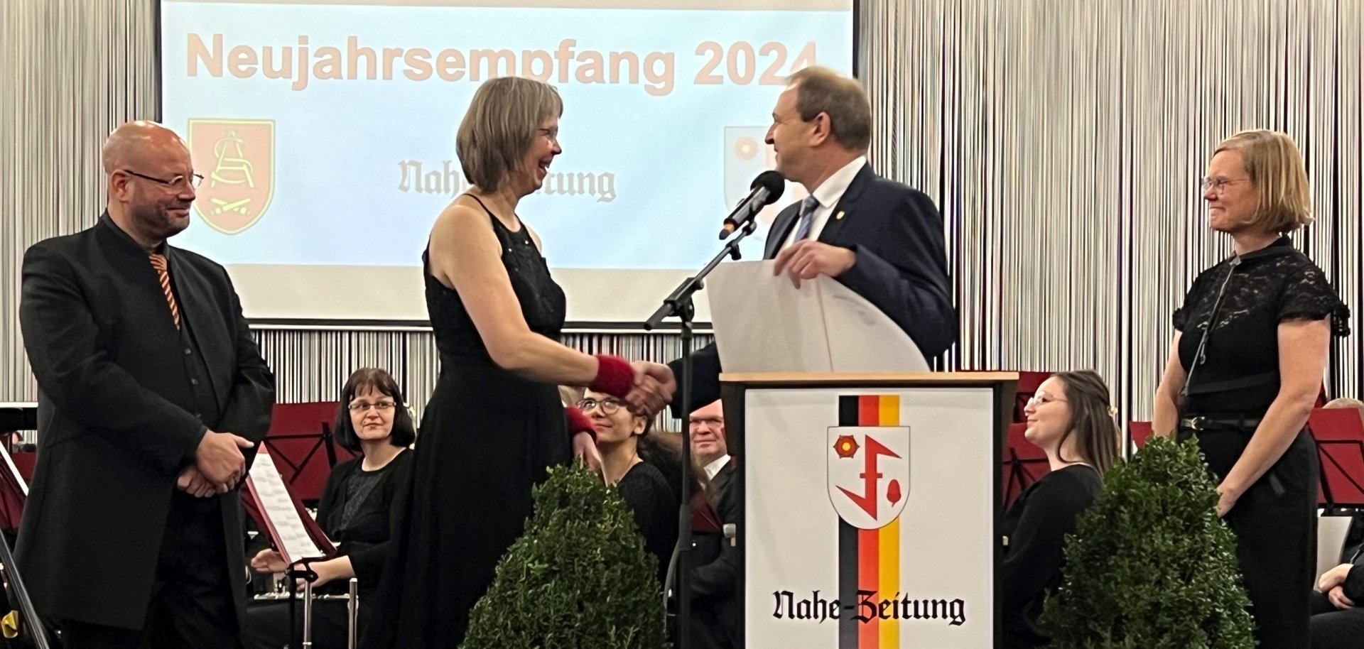 Das Foto zeigt die genannten Personen, die gemeinsam auf der Bühne des Neujahrsempfangs stehen. Der Oberbürgermeister gratuliert der Vorsitzenden und hält dabei die Urkunde in der Hand. Der Dirigent und die 2. Vorsitzende stehen links und rechts davon uns schauen zu. 