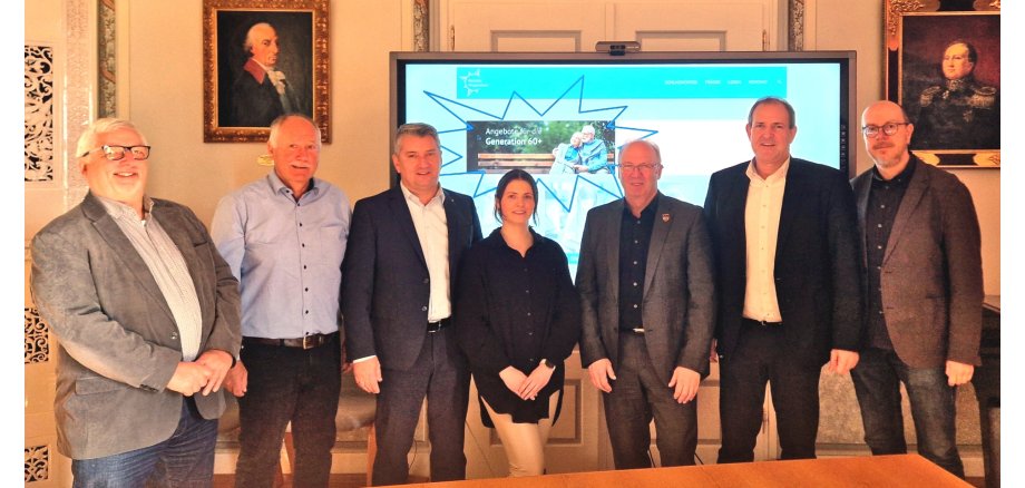 Das Foto zeigt die genannten Personen, die nebeneinander vor einem Bildschirm stehen und in die Kamera schauen. Auf dem Bildschirm ist die Internetseite mit dem Angebot der Seniorenplattform zu sehen.