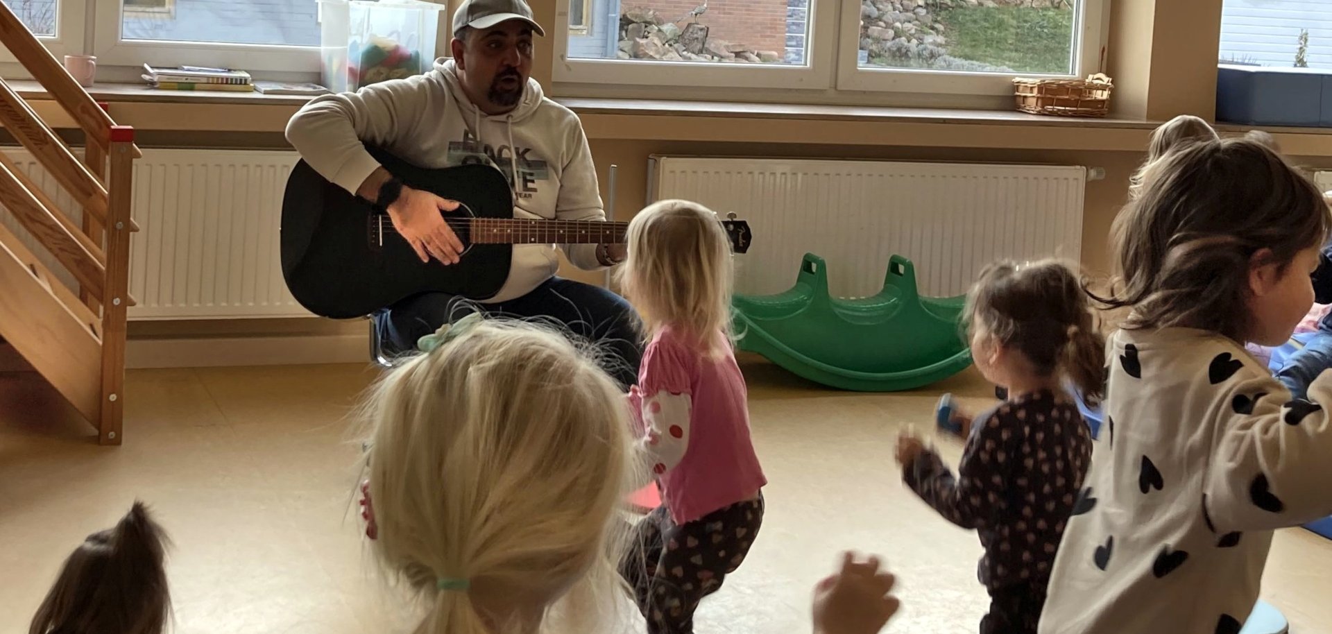 Das Foto zeigt einen Blick auf eine Übungsstunde in einem Kita-Raum. Im Hintergrund sitzt ein Musiklehrer mit Gitarre auf einem Stühl. im Vordergrund sind Kinder zu sehen, die sich zu der Musik bewegen.