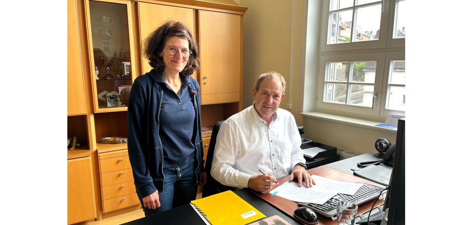 Das Foto zeigt OB Frühauf und Julia Besand. Der OB sitzt am Schreibtisch und unterzeichnet die Förderrichtlinie, Julia Besand steht daneben.