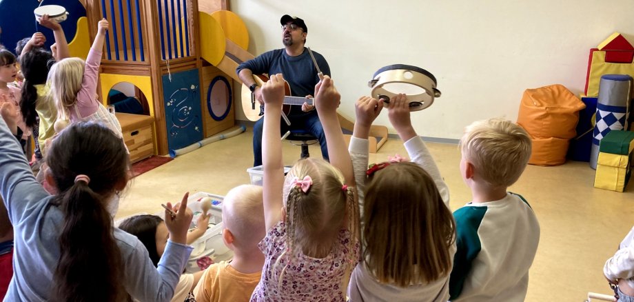 Das Foto zeigt den Blick in den Bewegungsraum während der Aktion "Musik-Mäuse". Im Hintergrund sitzt Mike Stauder mit einer Gitarre auf einem Stuhl und singt. Im Vordergrund tanzen die Kinder.