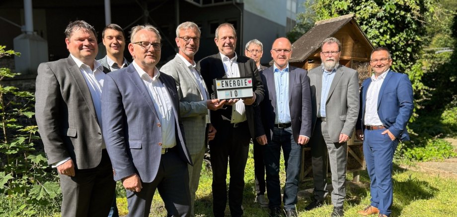 Das Foto zeigt die im Text genannten Personen. Sie stehen auf einer Wiese nebeneinander und schauen in die Kamera. Herr Johann und OB Frühauf halten ein Schild mit der Aufschrift Energie IO in den Händen.