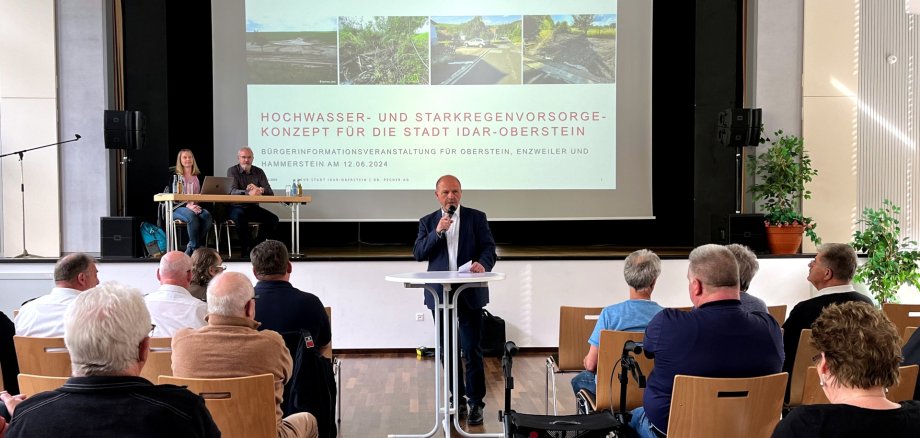 Das Foto zeigt einen Blick vom Zuschauerbereich zur Bühne. Vor der Bühne steht Bürgermeister Marx an einem Stehtisch und hält eine Ansprache. Auf der Bühne sitzen Herr Webler und Frau Baron an einem Tische neben der Leinwand, auf der die Präsentation zum HSVK zu sehen ist.