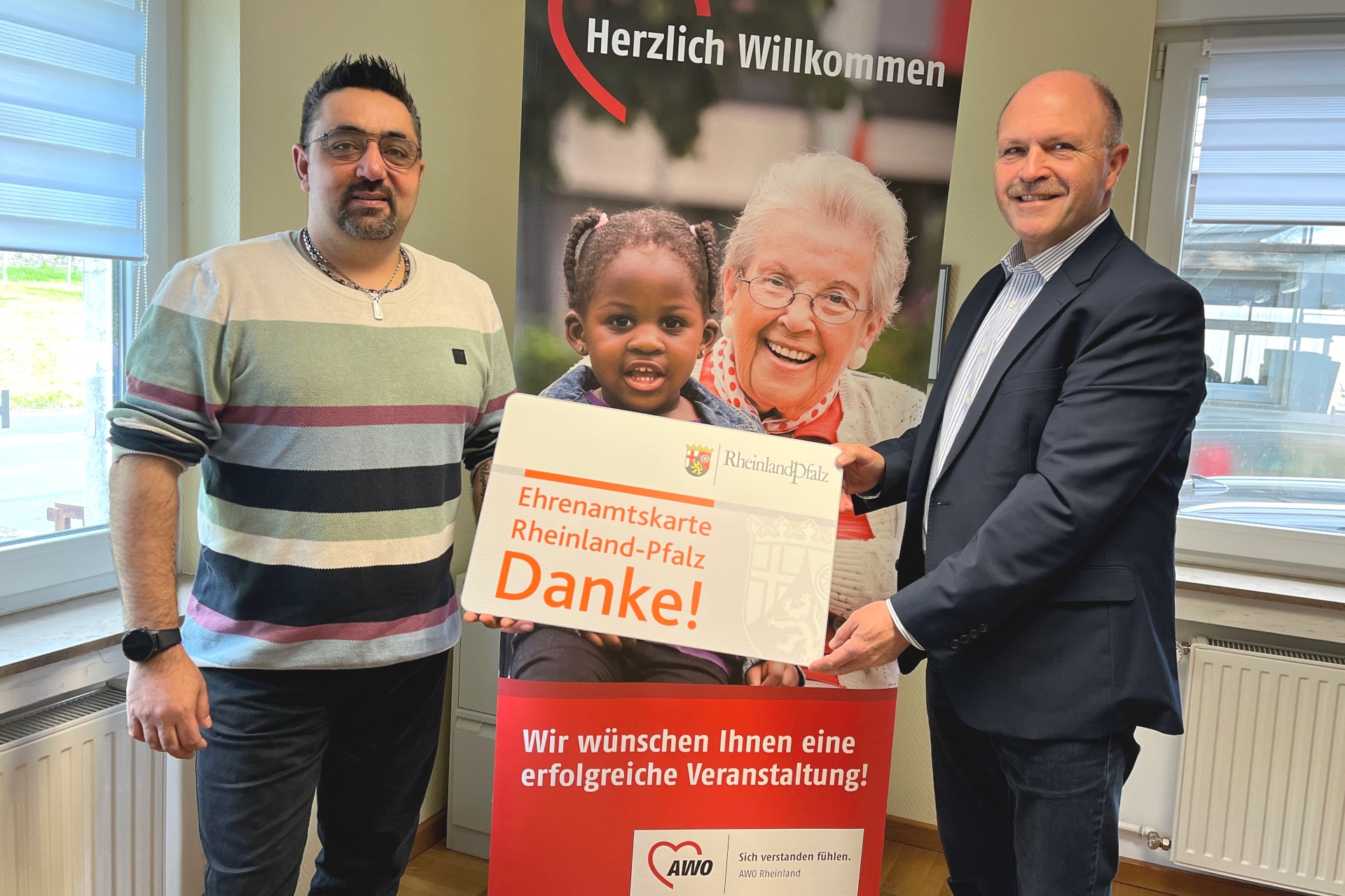 Das Foto zeigt die beiden genannten Personen. Sie halten gemeinsam eine überdimensionale Ehrenamtskarte in die Kamera, im Hintergrund ist ein Roll-Up der AWO zu sehen.