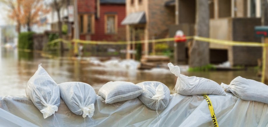 Das Foto zeigt im Vordergrund einen Damm aus Sandsäcken, im Hintergrund sind unscharf Häuser zu sehen, die im Hochwasser stehen und mit Trassierband abgesperrt sind.