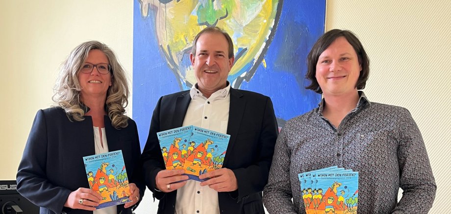 Das Foto zeigt die genannten Personen. Sie stehen nebeneinander, halten Ferienhefte in der Hand und schauen in die Kamera.
