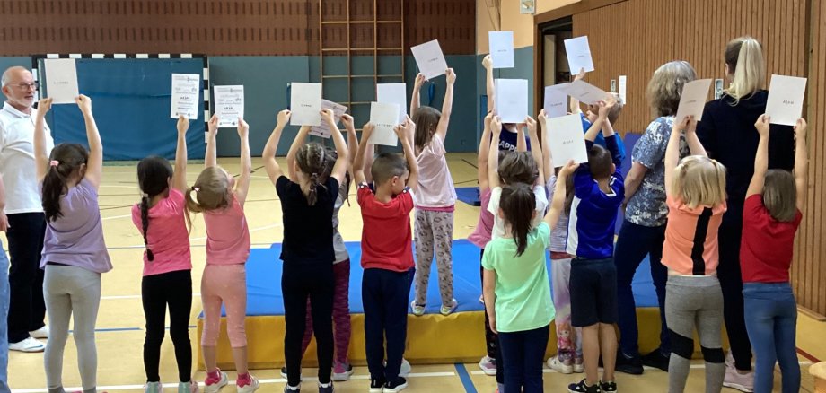 Das Foto zeigt viele Kinder, die in einer Turnhalle auf einer großen Matte stehen. Sie drehen der Kamera den Rücken zu und halten ihre Urkunden hoch.