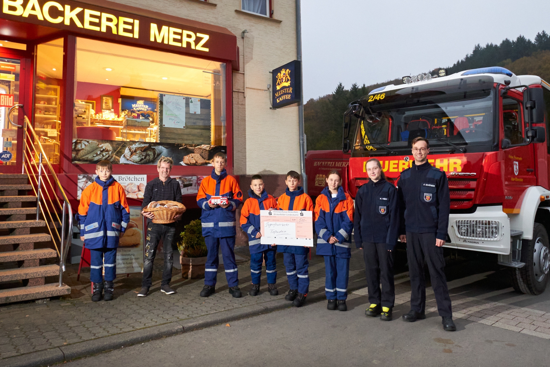 Das Foto zeigt eine Abordnung der Jugendfeuerwehr, die gemeinsam mit Bäckermeister Michael Merz vor dem Bäckerladen steht. Sie stehen nebeneinander, Bäcker Merz hält einen Korb mit Brezeln in der Hand, zwei Mitglieder der Feuerwehr einen großen Spendenscheck. Dahinter ist ein Feuerwehrauto zu sehen. 