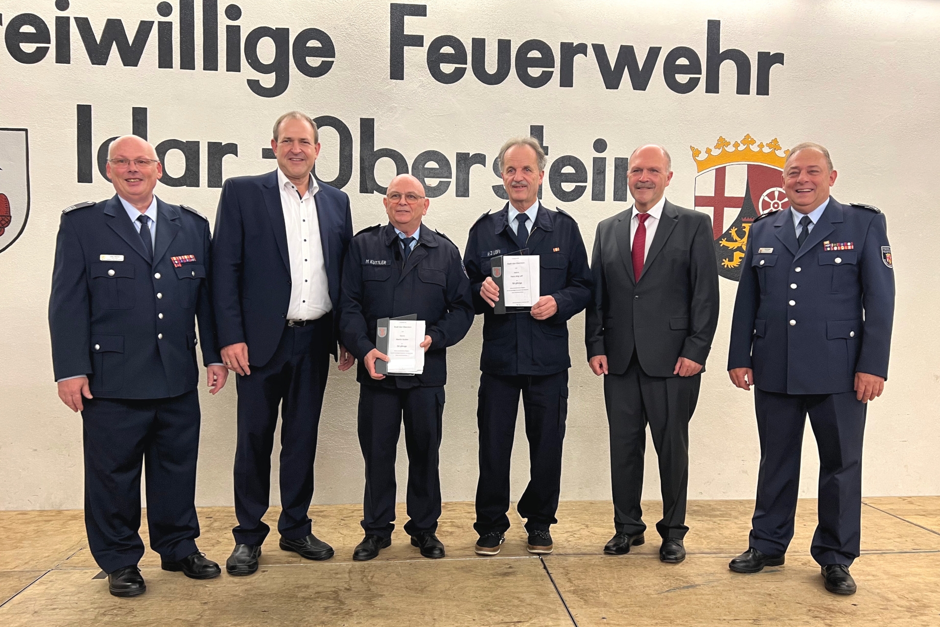 Das Foto zeigt die Geehrten mit Gratulanten. Sie stehen nebeneinander auf der Bühne und blicken in die Kamera.