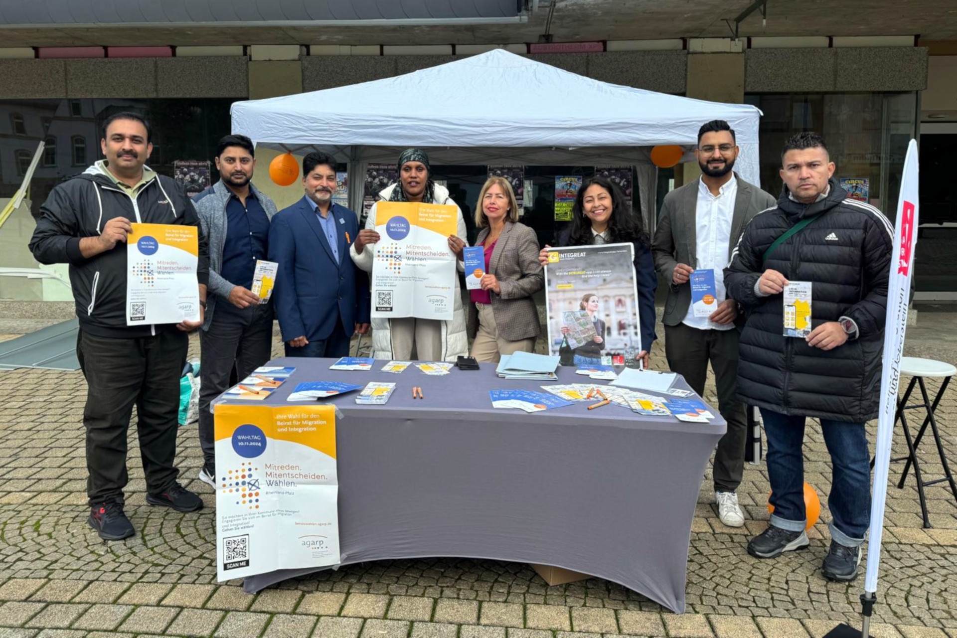 Das Foto zeigt den Informationsstand mit Flyern und Plakaten. An dem Stand stehen mehrere Personen, die ebenfalls Flyer und Plakate in den Händen halten und in die Kamera blicken.