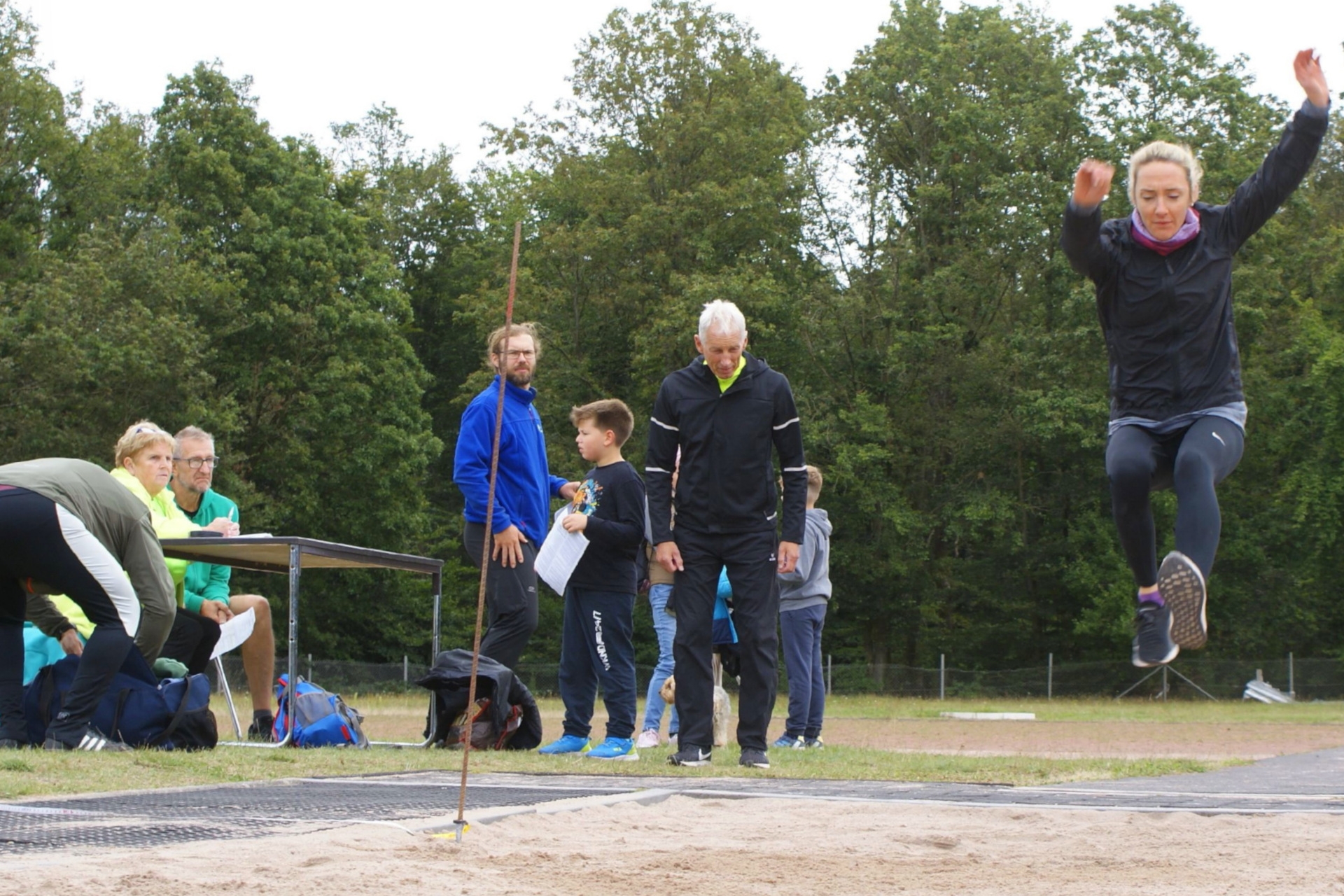 Das Foto zeigt eine Sportlerin, die Weitsprung macht und mehrere Abnehmer, die den Sprung beobachten bzw. am Tisch mit den Startkarten sitzen.