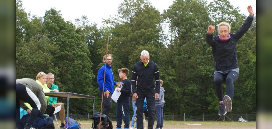 Das Foto zeigt eine Sportlerin, die Weitsprung macht und mehrere Abnehmer, die den Sprung beobachten bzw. am Tisch mit den Startkarten sitzen.
