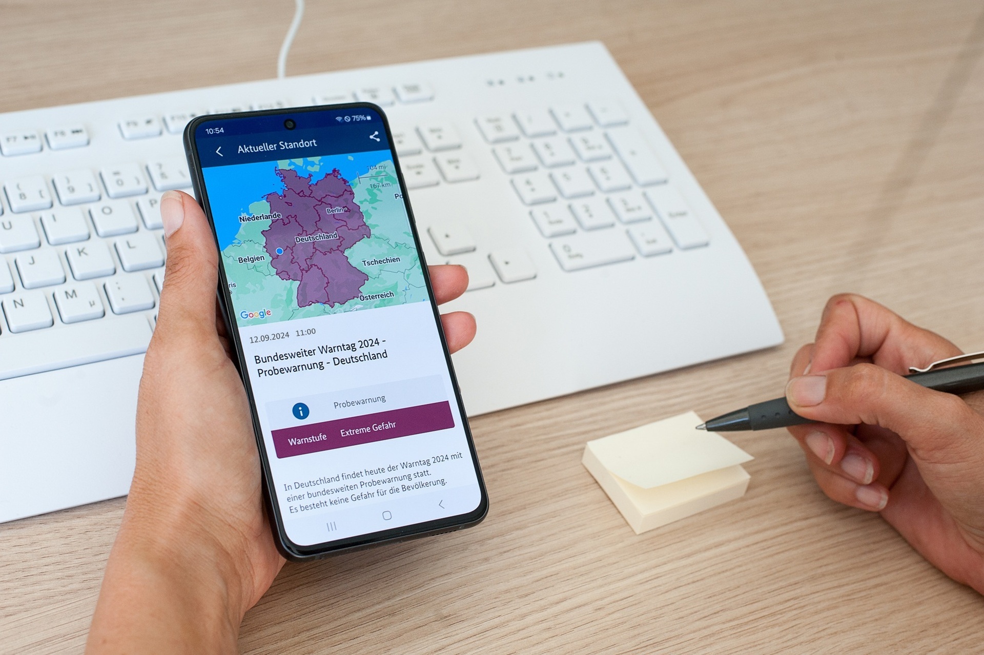 Das Foto zeigt eine Hand, die ein Handy hält. auf dem Handy ist die Warn-APP NINA mit einer Probewarnung zu sehen.