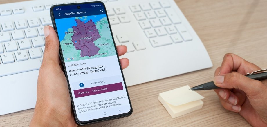 Das Foto zeigt eine Hand, die ein Handy hält. auf dem Handy ist die Warn-APP NINA mit einer Probewarnung zu sehen.