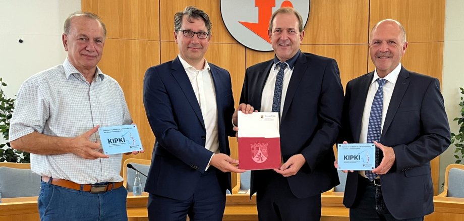 Das Foto zeigt die genannten Personen. Sie stehen im Sitzungssaal nebeneinander und blicken in die Kamera. Staatssekretär und Oberbürgermeister halten gemeinsam den Förderbescheid, Landtagsabgeordneter und Bürgermeister halten jeweils ein KIPKI-Schild.