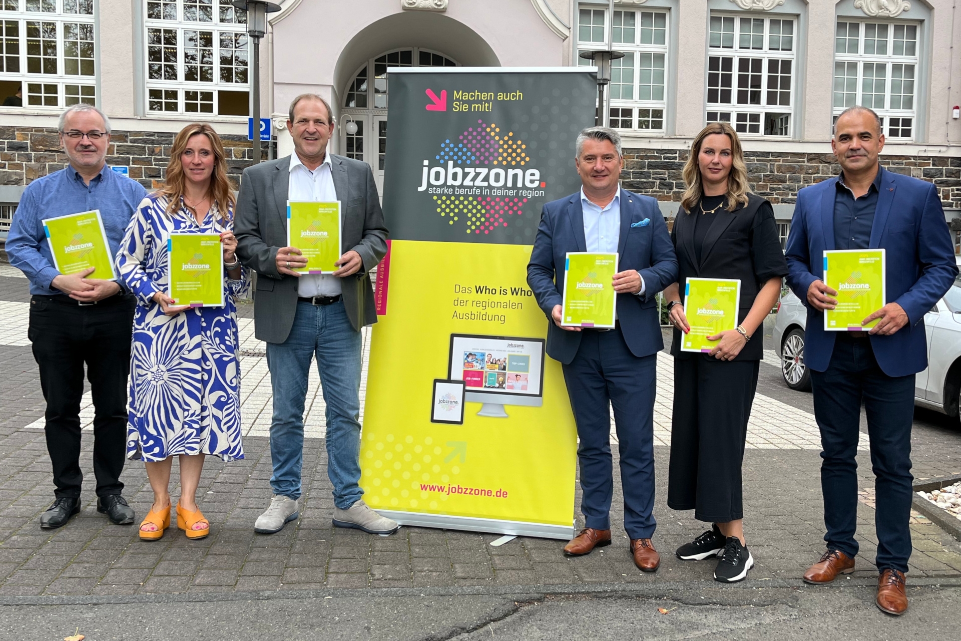 Das Foto zeigt die genannten Personen, die nebeneinander vor der Fassade des Stadthauses stehen und in die Kamera blicken. Jede Person hält eine "jobzzone" in der Hand. In der Mitte steht ein Rollup Display mit der Aufschrift "jobzzone".