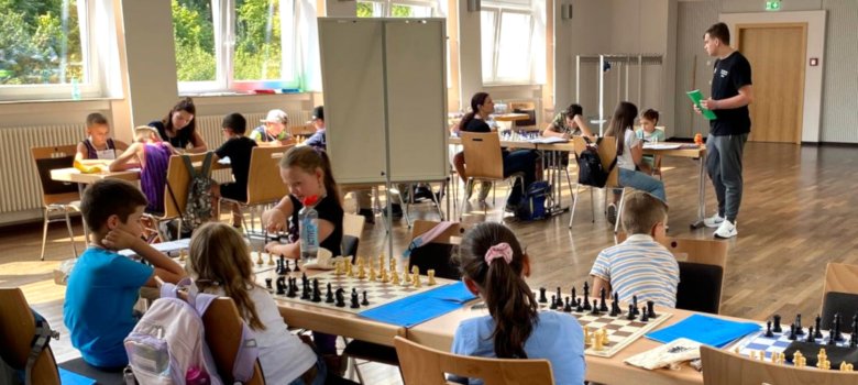 Das Foto zeigt einen Blick in die Göttenbach-Aula. An mehreren Tischen sitzt jeweils ein Kind. Auf dem Tisch liegt ein Schachbrett mit Figuren. Im Hintergrund steht ein Trainer und erklärt den Kindern etwas.