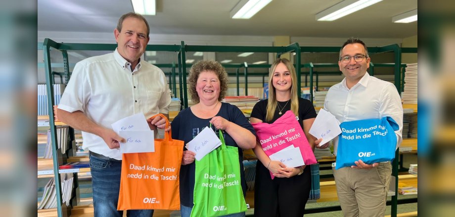 Das Foto zeigt vier Personen, die nebeneinander vor einem Regal mit Schulbüchern stehen. Sie halten die bunten OIE-Stofftaschen in den Händen und schauen in die Kamera.
