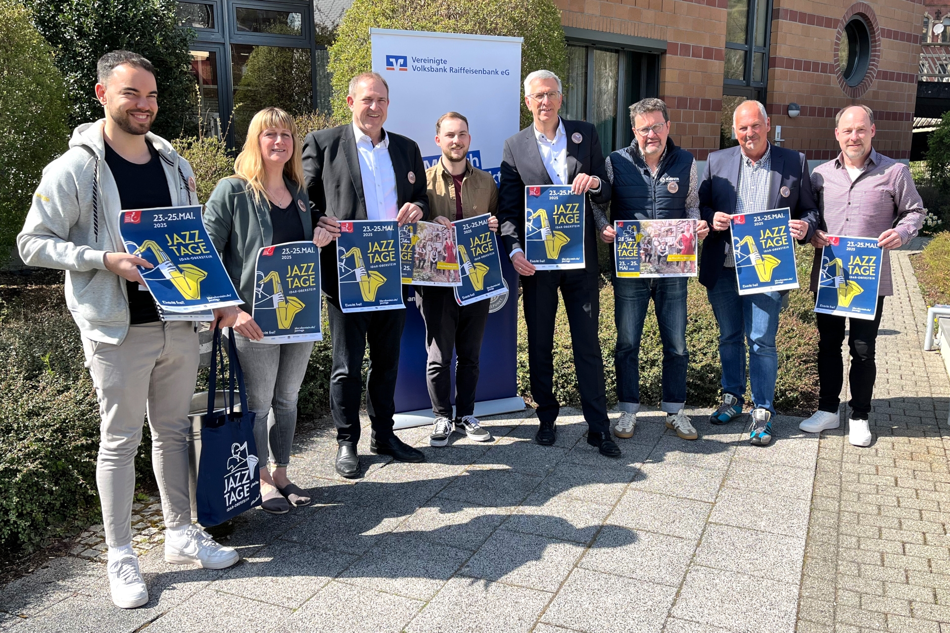 Das Foto zeigt 8 Personen, die im Freien nebeneinander stehen und Plakate der Jazztage in den Händer halten.