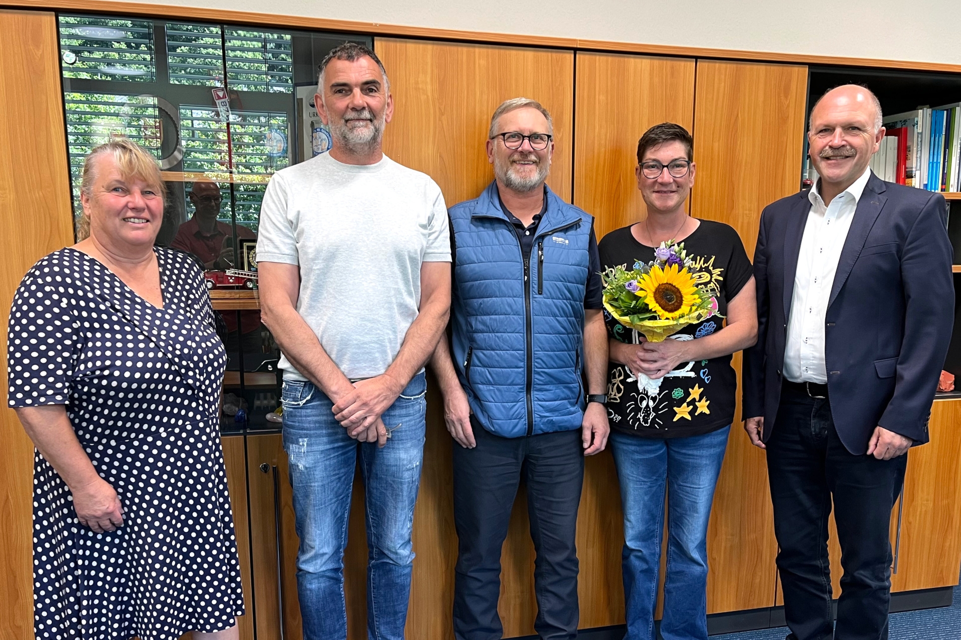 Das Foto zeigt die genannten Personen, die nebeneinander vor einer Schrankwand stehen und in die Kamera schauen. Nicole Hermann hält einen Blumenstrauß in den Händen.