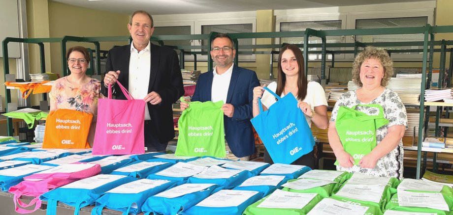Das Foto zeigt die genannten Personen. Sie stehen nebeneinander hinter einem Tisch mit Stapeln von gefüllten Taschen. Die Personen halten jeweils eine Tasche in der Hand und schauen in die Kamera.