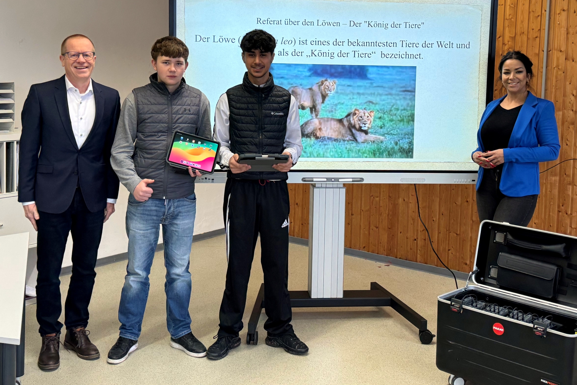Das Foto zeigt die beiden genannten Personen, die gemeinsam mit zwei Schülern vor einem Whiteboard stehen. Die Schüler haben noch jeweils ein Tablet in der Hand.