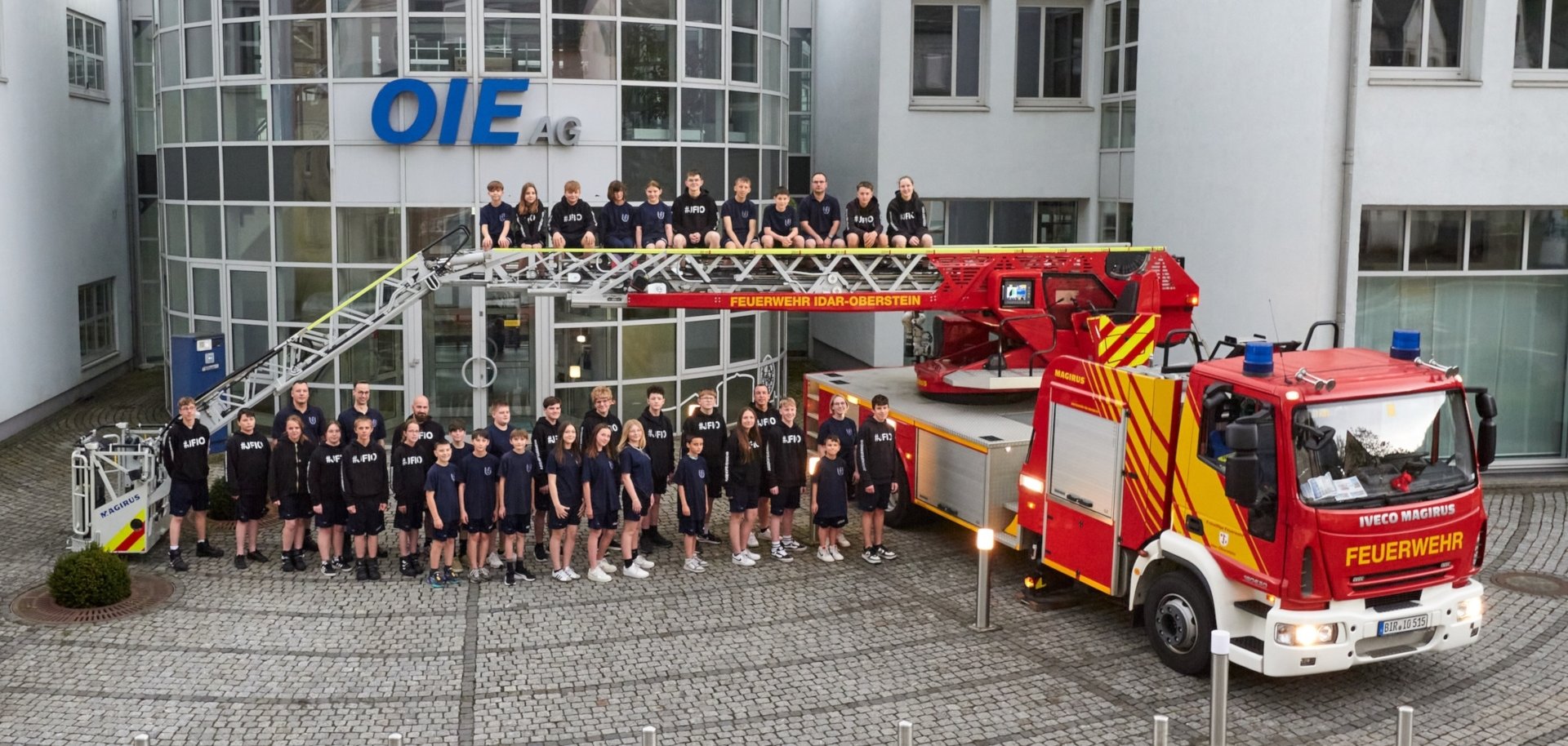 Das Foto zeigt die Mitglieder der Jugendfeuerwehr Idar-Oberstein. Sie stehen in den neuen Outfits vor dem Verwaltungsgebäude der OIE. Zu sehen ist auch die Drehleiter der Feuerwehr. Einige Mitglieder sitzen auf der ausgefahrenen Leiter.