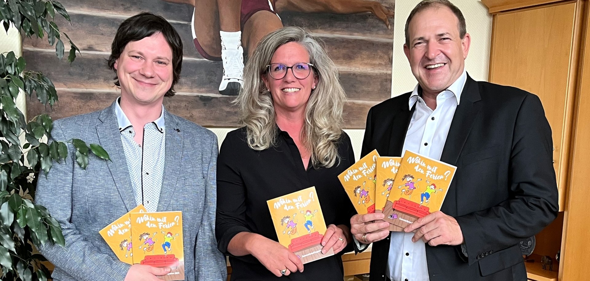 Das Foto zeigt die genannten Personen. Sie stehen im Büro des Oberbürgermeisters nebeneinander, blicken in die Kamera und halten jeweils ein Ferienheft in der Hand.