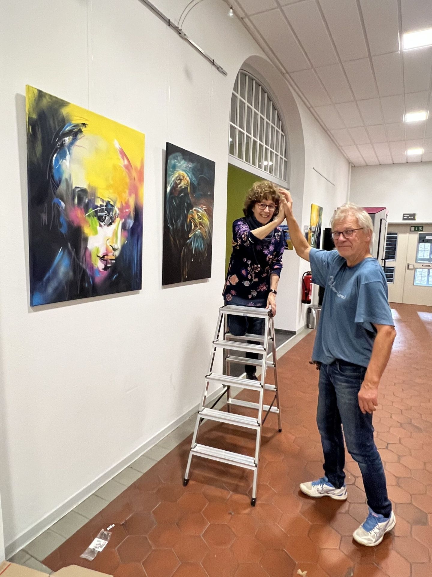 Das Foto zeigt Carmen Kroese auf der Leiter mit ihrem Helfer vor zwei Bildern im Foyer. 