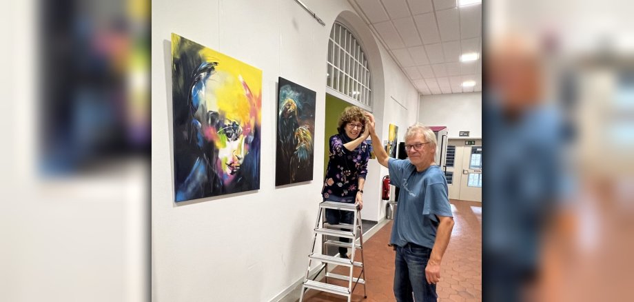 Das Foto zeigt Carmen Kroese auf der Leiter mit ihrem Helfer vor zwei Bildern im Foyer. 