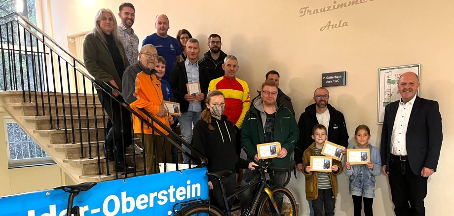Leider konnten nicht alle Gewinner an der Preisverleihung teilnehmen, aber ein Großteil versammelte sich zum abschließenden Gruppenfoto. Das Foto zeigt die Preisträger, die auf dem Treppenaufgang zur Göttenbach-Aula stehen. Am Geländer hängt ein Banner des STADTRADELN, davor steht ein Fahrrad.