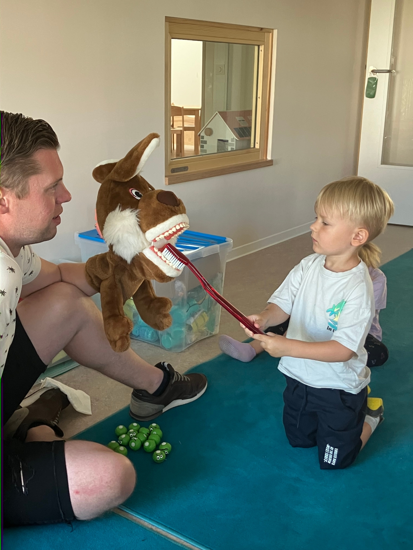 Das Foto zeigt den Zahnarzt, der eine Handpuppe mit geöffnetem Mund hält. Davor sitze ein Junge und putzt die Zähne der Handpuppe mit einer überdimensionalen Zahnbürste.