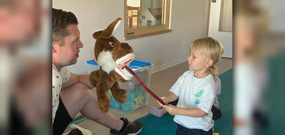 Das Foto zeigt den Zahnarzt, der eine Handpuppe mit geöffnetem Mund hält. Davor sitze ein Junge und putzt die Zähne der Handpuppe mit einer überdimensionalen Zahnbürste.