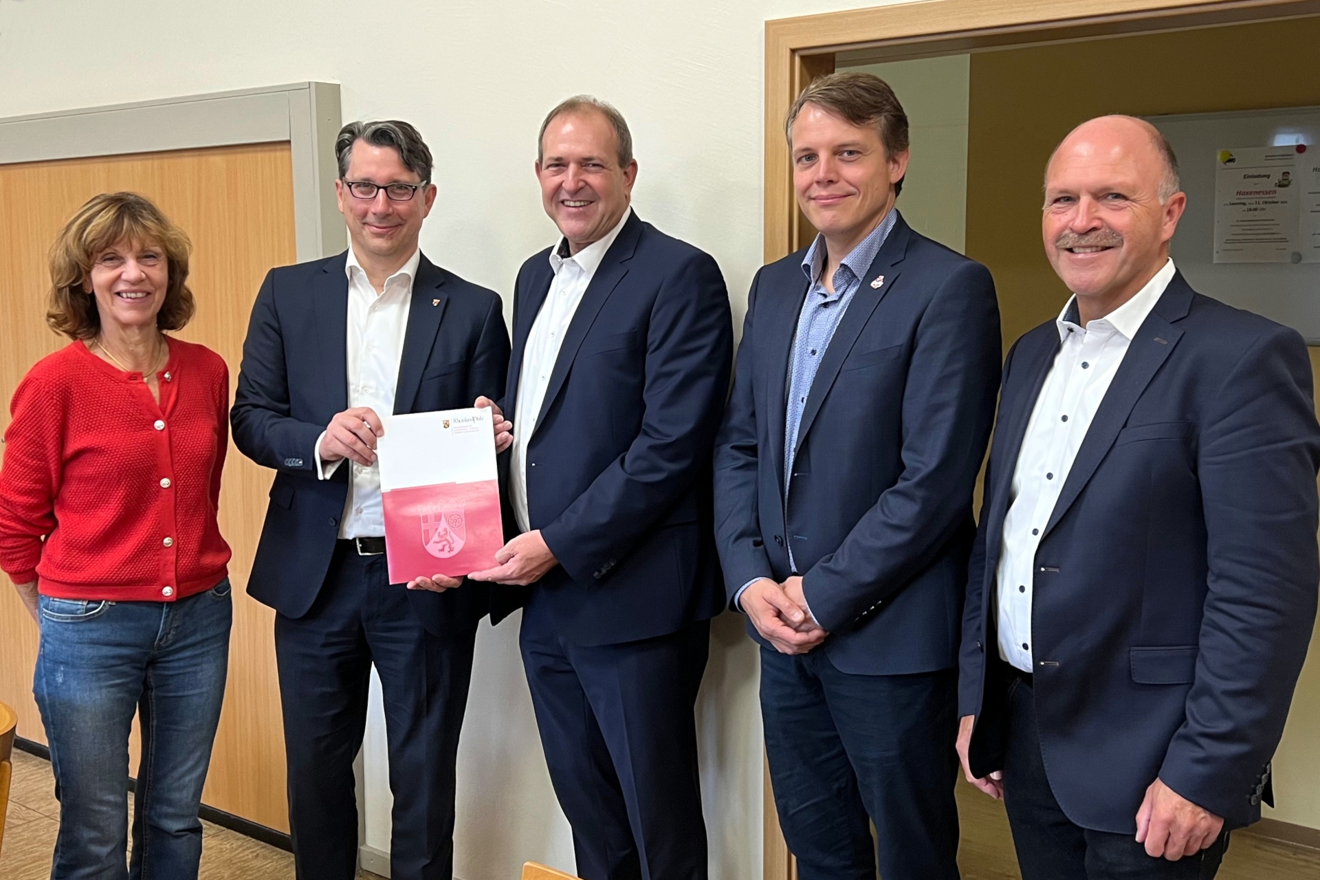 Das Foto zeigt die genannten Personen. Sie stehen nebeneinander in einem Raum der Kita. Der Staatssekretär überreicht dem Oberbürgermeister den Förderbescheid.