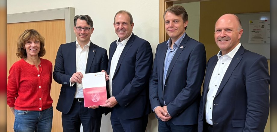 Das Foto zeigt die genannten Personen. Sie stehen nebeneinander in einem Raum der Kita. Der Staatssekretär überreicht dem Oberbürgermeister den Förderbescheid.