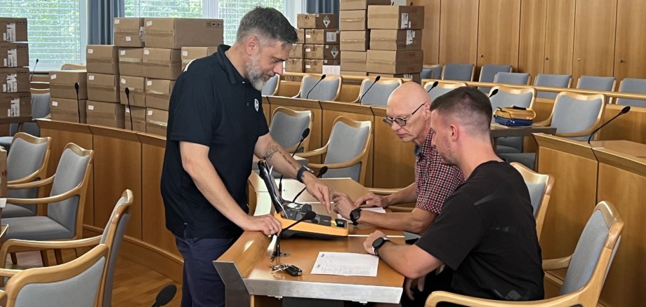 Übergabe von Defibrillatoren an Michael Brill für die Stadenhalle und Sven Dalheimer für die Polizeiinspektion durch Oliver Köppl von der Herstellerfirma ultraMEDIC. Das Foto zeigt zwei Männer, die von einem dritten Mann eine Einweisung in die neuen Defibrillatoren bekommen.